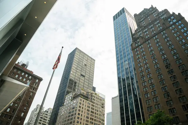 Manhattan şehir merkezindeki yüksek binaların önünde bir Amerikan bayrağı yükseliyor. Cam, tuğla ve taş mimari çeşitli şehirleri temsil eder..