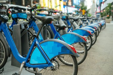 Yoğun bir şehir kaldırımındaki bir rıhtım istasyonunda bir sıra mavi kiralık bisiklet düzgünce dizildi. Arka planda bulanık trafik, ışıklar ve yüksek binalar görülüyor. Bu da canlı bir kentsel çevre olduğunu gösteriyor..