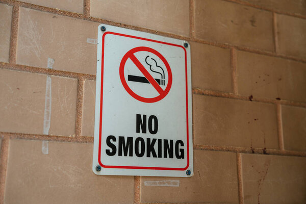 No smoking sign prominently displayed on a brick wall in an outdoor setting. Bright red circle with diagonal line through cigarette conveys cautionary message clearly.