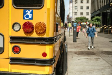 Şehir caddesinde park etmiş parlak sarı okul otobüsü erişilebilirlik sembolü içeriyor. Kentsel çevre güneşli gökyüzü altında sakin bir atmosfer sergiliyor, ulaşım erişilebilirliğini vurguluyor.