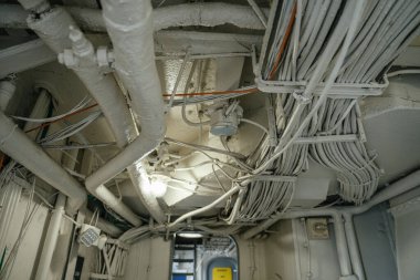 Askeri bir geminin tavanına yakından bakınca yoğun bir boru ve kablo şebekesi görülüyor. Sahne, deniz mühendisliğinin karmaşık altyapısı ve endüstriyel tasarımını vurguluyor..