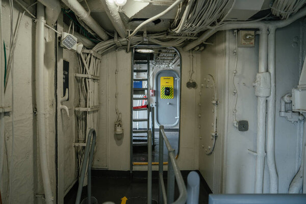 A narrow corridor inside a military ship shows exposed cables, metal pipes, and a yellow safety sign on a door in the background. The industrial space is dimly lit and utilitarian in design.