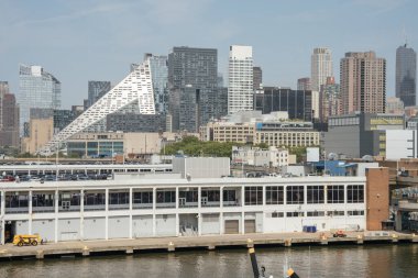 Modern bir üçgen bina New York şehrinin gökdelenleri arasında göze çarpıyor. Ön planda Hudson Nehri yakınlarında bir liman deposu ve inşaat araçları görünüyor..