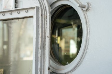 Yıpranmış bir gemi kapısı ve yuvarlak bir pencere. Cam yüzeyde ışığın ve bulanık çevrenin yansımaları görülebilir..