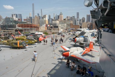 New York City, New York, ABD - 07.30. 2024: Bir uçak gemisinin güvertesinde birden fazla klasik askeri helikopter ve uçak sergilenmektedir. İnsanlar etrafta dolaşıp arka planda Manhattan silueti olan uçakları inceliyor..