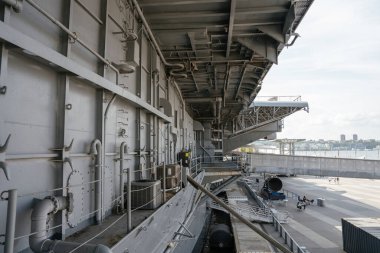 Uçak gemilerinin yan tarafında görünür borular, demirleme ipleri ve yapısal kirişler bulunur. Aşağıda, rıhtım nehir kenarındaki banklar ve ziyaretçilerle halka açık bir yürüyüş yoluna ev sahipliği yapıyor..