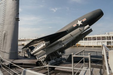 Bir liman müzesinde korunmuş bir Amerikan denizaltısının güvertesinde büyük bir donanma füzesi sergileniyor. Denizaltı açık mavi gökyüzünün altında bir şehir limanının yanında demirli..