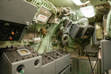 Elektronik paneller, kablolar, anahtarlar ve analog ekranlarla dolu karmaşık bir denizaltı. Bir uyarı işareti ve teknik aletler Soğuk Savaş dönemi sistemlerini vurguluyor.