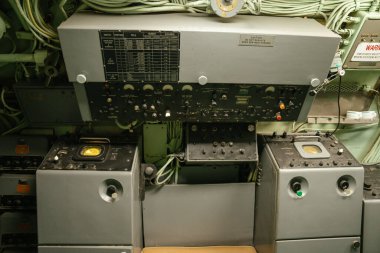 Bir donanma denizaltısının içindeki radar ve sinyal kontrol ünitesinin yakın görüntüsü. Soğuk Savaş döneminin korunan panelini analog araçlar, kablolar ve uyarı etiketleri kapsıyor.