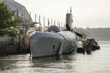 Bir askeri denizaltı nehir kıyısındaki bir rıhtıma demirlemiş, Amerikan bayrakları ve yollarıyla dolu. Ağaçlar ve müze binaları arka planı çerçevelerken, metalik yüzey gün ışığını yansıtıyor.