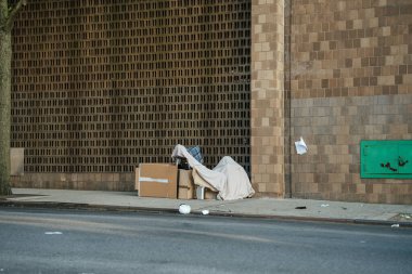 Mukavva ve kumaştan yapılmış derme çatma bir barınak şehrin kaldırımında tuğla bir duvara karşı duruyor. Sahne, kentsel evsizliği ve metropoliten bir ortamda temel yaşam koşulları için mücadeleyi yansıtıyor..