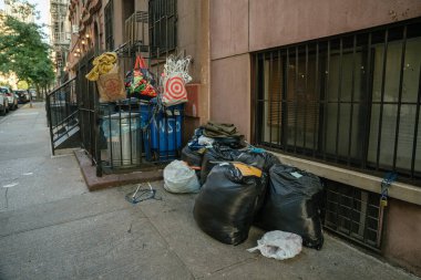 New York City, New York, ABD - 07.30. 2024: Siyah çöp torbaları ve geri dönüşüm kutuları kapılı bir merdiven boşluğunun yanındaki iskan kaldırımı boyunca yığılmıştır. Çeşitli çantalar, askılar ve dağınıklık kumlu bir sokak sahnesi oluşturur..