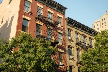 Yangın merdivenleri olan klasik New York apartmanlarının yakın görüntüsü. Kırmızı ve bej tuğlalı cepheler Manhattan 'daki yeşil ağaçların üzerinde altın güneş ışığında duruyor..