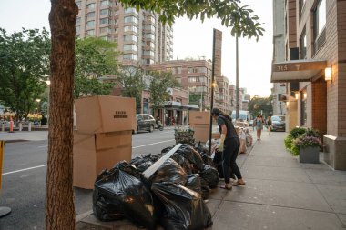 New York City, New York, ABD - 07.30. 2024: Bir temizlik işçisi gün batımında kaldırımda siyah çöp torbalarını ayırıyor. Yoğun şehir caddesinde büyük karton kutular ve geri dönüşüm çantaları görülebiliyor.
