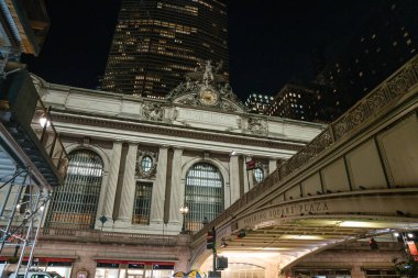 New York City, New York, ABD - 07.30. 2024: Grand Central Terminali Manhattan şehir merkezindeki yüksek gökdelenlerin altında gece aydınlatılmaktadır. Pershing Square Plaza köprüsü ve yakındaki iskeleler dinamik şehir atmosferine katkı sağlıyor..