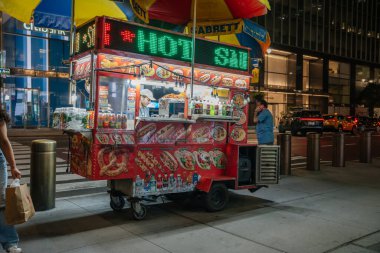 New York City, New York, ABD - 07.30. 2024: Renkli bir sosisli sandviç büfesi gece New York 'ta işlek bir cadde köşesinde yemek servisi yapıyor. Satıcı, işaretler, soslar ve şehir trafiğiyle çevrili parlak ışıkların altında atıştırmalıklar hazırlar..