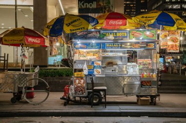 New York City, New York, ABD - 07.30. 2024: Parlak şemsiyeler ve ışıklı tabelayla süslenmiş canlı bir helal gıda arabası gece kaldırımına park edilmiştir. Arabada sosisli sandviçler, jiroskoplar ve içkiler var. Yoldan geçenleri geç saatlere kadar davet ediyorlar.