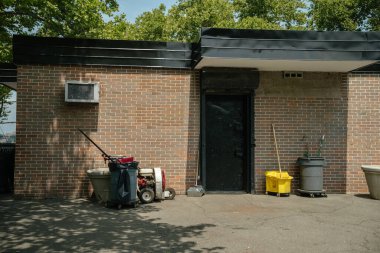 Siyah metal kapılı bir tuğla bina temizlik malzemeleri ve çöp kutularıyla çevrilidir. Parlak güneş ışığı altında bir süpürge kovası, süpürge ve bir yaprak üfleme arabası görünür..