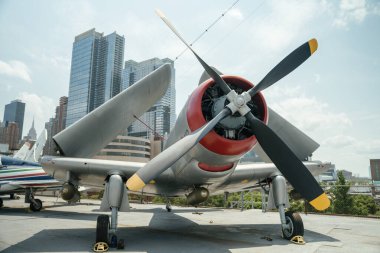 Katlanmış kanatları ve kırmızı motor detayı olan gümüş renkli bir pervane uçağının ön görüntüsü. Uçak, modern bir şehir siluetine karşı açık havada sergileniyor..