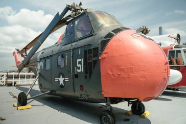51 numaralı gri ve kırmızı askeri helikopter ve Amerikan donanması işaretleri bir uçak gemisinde sergileniyor. Helikopter açık havada bir müzede güneşli bir gökyüzünün altında duruyor..