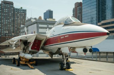 Görünür kokpiti, füzeleri ve iniş takımları olan beyaz ve kırmızı bir donanma savaş uçağının ön ve yan görüntüsü. Uçak, şehir binalarıyla çevrili bir müze güvertesinde park halinde..