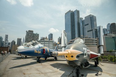 New York City, New York, ABD - 07.30. 2024: İki klasik donanma savaş jeti modern gökdelenlerin arkasında yükseldiği bir çatı güvertesinde sergileniyor. Jetler arasında bir Mavi Melek uçağı ve 210 işaretli koyu gri bir model var..