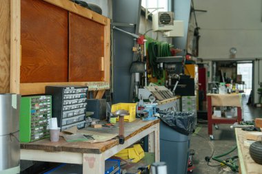 Küçük parçalar, çekmeceler ve aletlerle dolu düzenli bir tezgah restorasyon hangarında kuruluyor. Düzenli çalışma alanının arka planını ekipman, hortum ve el arabaları doldurur.