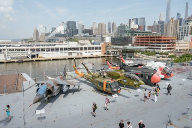 New York City, New York, ABD - 07.30. 2024: Tarihi bir uçak gemisi müzesinin güvertesinde askeri uçak ve helikopter sıralaması sergilenmektedir. Ziyaretçiler, uçağın üzerinde bir şehir silueti ve kıyı şeridi bulunan yürüyüş ve gözlem yapıyorlar.