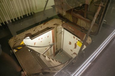 Yıpranmış bir denizaltı kapağından aşağıya inen dik metal bir merdiven. Kırık boya, uyarı işareti ve açıkta duran borular endüstriyel bir his veriyor..