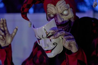 Kırmızı ve siyah giyen ürkütücü soytarı mavi gece lambasının altında beyaz bir palyaço maskesi tutuyor. Parlayan gözler, keskin dişler ve dramatik kostüm ürkütücü bir Cadılar Bayramı sahnesi oluşturur..