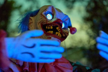 Ürkütücü palyaço mavi ışıklı elleri ve hipnotik spiral gözleriyle öne uzanıyor. Kırmızı burun, sarı dişler ve dramatik ışıklandırma korkunç bir Cadılar Bayramı sahnesi yaratır..