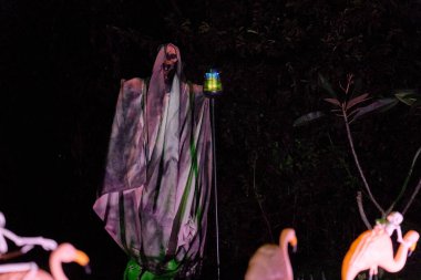 Kapüşonlu Azrail iskeleti karanlık bir cadılar bayramı vitrininde parlayan bir fener tutuyor. Ürkütücü pelerinli figür geceleri bahçe süslemelerinin arasında duruyor..