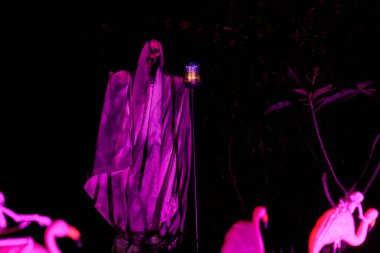 Fenerli iki uzun kukuletalı orakçı ürkütücü bir çimen sahnesinde mor parlıyor. Gece vakti perili ev bahçesi ürkütücü ışıklar ve gölgelerle doludur..