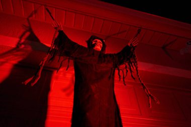 Kukuletalı, yırtık pırtık elbiseli bir iskelet güçlü kırmızı ışıkta yükselen kollarıyla asılı duruyor. Geceleri dramatik gölgelerle dolu ürkütücü Cadılar Bayramı bahçesi dekorasyonu.