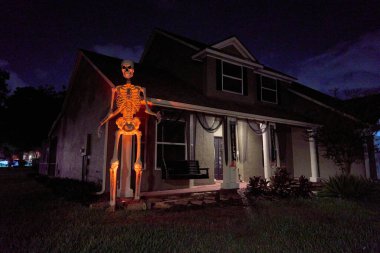 Yükselen iskelet figürü, kolonları olan bir verandanın yanında turuncu parlar. Karanlık çöktükten sonra banliyö evinde ürkütücü bir cadılar bayramı gösterisi..