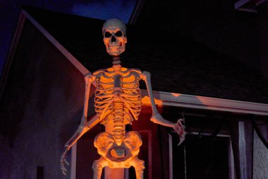 Yüksek bir iskelet figürü turuncu spot ışıkları altında bir evin verandasında duruyor. Karanlık sahneye kemikleri ve kafatası olan ürkütücü cadılar bayramı dekoru hakim..