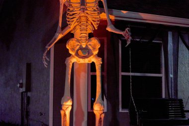 Yüksek bir iskelet dekorasyonunun veranda sütunuyla parlayan turuncu rengini yakından görebiliriz. Geceleri arkada kemikler ve pencerelerle ürkütücü cadılar bayramı görüntüsü.