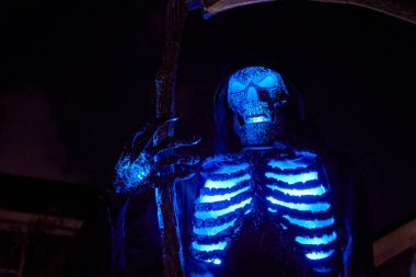 Kapüşonlu kafatası figürü geceleri parlak kaburgaları ve gözleri ile mavi parlar. Dramatik ışıklandırma ve derin gölgelerle ürkütücü Cadılar Bayramı dekorasyonu.