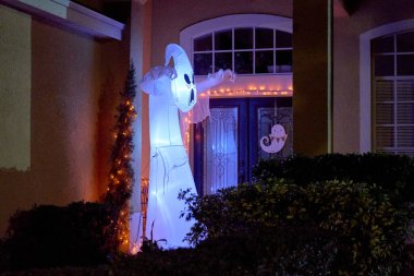 Uzun, beyaz, şişme bir hayalet geceleri turuncu ışıklarla ön kapının yanında parlıyor. Cadılar Bayramı bahçe dekorasyonları banliyödeki bir eve ürkütücü, şenlikli bir giriş yaratır..