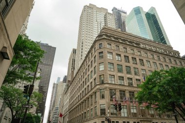 Amerikan bayraklı tarihi bir bina 5. Cadde 'nin köşesinde duruyor. Etrafı uzun ve modern gökdelenlerle çevrili. Sokak manzarası bulutlu bir gökyüzü altında eski ve yeni mimari tarzları harmanlıyor.