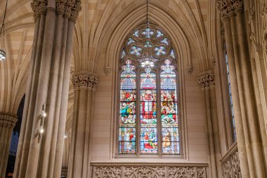 Görüntü, New York 'taki St. Patricks Katedrali' nin içindeki İncil sahnelerini gösteren canlı bir vitraylı pencereyi gösteriyor. Uzun taş sütunlar ve karmaşık Gotik kemerlerle çevrelenmiş sanat eserleri kutsallığa güzellik ve ruhani sembolizm katıyor.