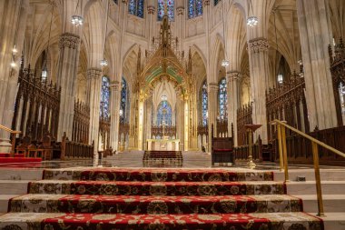 Resim, New York 'taki St. Patricks Katedrali' nin altın Gotik kemerler ve vitray pencerelerle çerçevelenmiş büyük mihrabını yakalıyor. Süslü sütunlar, oyma ahşap işçiler ve kutsal mabede kadar uzanan zengin kırmızı halılar..