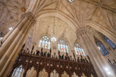Fotoğraf, New York 'taki St. Patricks Katedrali' ndeki tırtıklı tavan, detaylı koro ekranı ve vitraylı pencereleri vurguluyor. Süslü sütunlar ve sıcak ışıklandırma Gotik Diriliş mimarisinin güzelliğini arttırır..