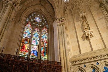 Bu resim, Gotik kemerli bir çerçeve içinde geçen İncilsel sahneleri gösteren renkli bir vitray penceresi sergiliyor. Sağda, bir azizin taş heykeli katedrallerin üzerine yerleştirilmiş..