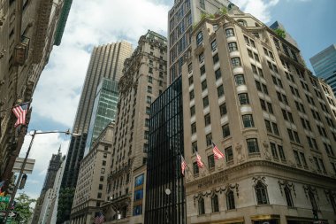 New York 'taki Beşinci Cadde üzerinde tarihi ve modern gökdelenlerin bir karışımı yükseliyor ve önünde Amerikan bayrakları sallanıyor. Sahne, güneşli bir günde mimari kontrastı ve vatansever şehir atmosferini yakalıyor..