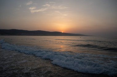 Su yüzeyinde küçük dalgalar var. Ufukta turuncu gökyüzünde kocaman yuvarlak bir güneş. Altın güneş ışınları su yüzeyine yansıyor.