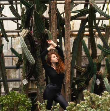 Bir kadın kaktüs bahçesinin önünde poz veriyor. O siyah bir gömlek ve siyah pantolon giyiyor.