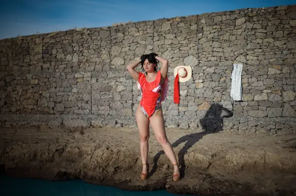 A woman in a red bikini stands in front of a stone wall. She is holding a straw hat and a red scarf. The scene is casual and relaxed, as the woman is dressed for a beach day.