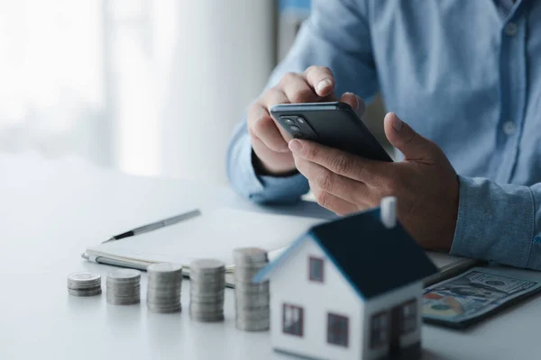 Person holding phone with house model and stack of coins calculating and recording income and expenses for financial planning, financial planning ideas to buy a house and real estate.
