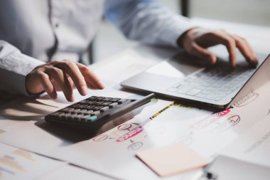 Businessman is using a calculator to calculate company financial figures from earnings papers, a businessman sitting in his office where the company financial chart is placed.